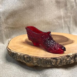 May include: A translucent ruby red glass shoe figurine featuring a detailed pressed pattern of stars and geometric shapes across the exterior surface, including a small raised heel and a pointed toe design, crafted from molded glass material.