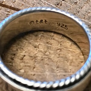 Op de afbeelding: Close-up van een zilveren ring met een getextureerd, touwachtig ontwerp rond de randen. De binnenkant van de ring is gestempeld met "@t&t" en "925". De ring ligt op een houten oppervlak.