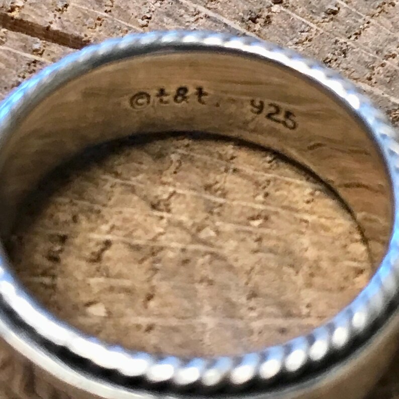 Op de afbeelding: Close-up van een zilveren ring met een getextureerd, touwachtig ontwerp rond de randen. De binnenkant van de ring is gestempeld met "@t&t" en "925". De ring ligt op een houten oppervlak.