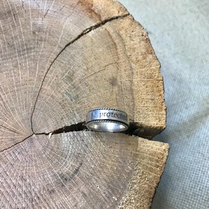 Op de afbeelding: Een zilveren ring met het woord "protected" gegraveerd op de band. De ring heeft een gevlochten touwontwerp rond de boven- en onderranden. De ring ligt op een houten oppervlak met textuur.