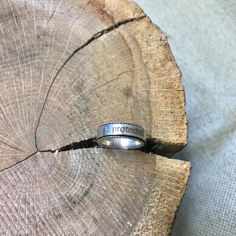 Op de afbeelding: Een zilveren ring met het woord "protected" gegraveerd op de band. De ring heeft een gevlochten touwontwerp rond de boven- en onderranden. De ring ligt op een houten oppervlak met textuur.
