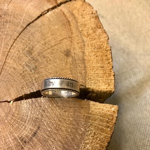 Op de afbeelding: Een zilveren ring met de woorden "by an" gegraveerd op de band. De ring heeft een touwachtig ontwerp rond de randen en ligt op een houten oppervlak. De ring is gemarkeerd met "MADE 925".