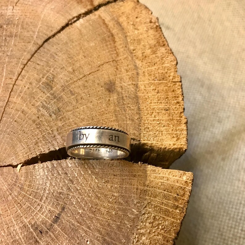 Op de afbeelding: Een zilveren ring met de woorden "by an" gegraveerd op de band. De ring heeft een touwachtig ontwerp rond de randen en ligt op een houten oppervlak. De ring is gemarkeerd met "MADE 925".