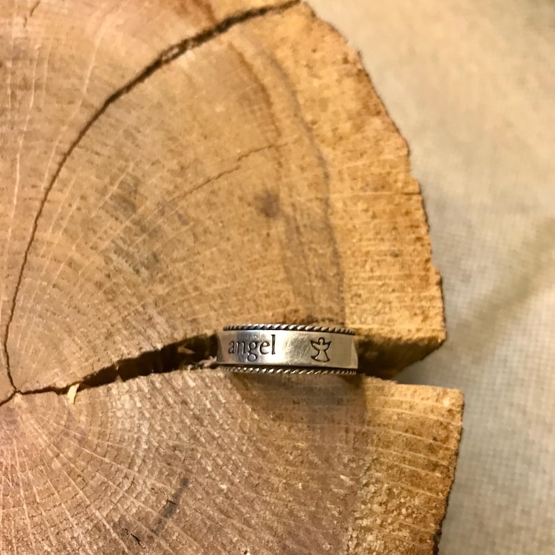 Op de afbeelding: Een zilveren ring met het woord "engel" en een kleine engelafbeelding. De ring is geplaatst op een rustieke houten achtergrond, die het ontwerp en de details van de ring benadrukt.