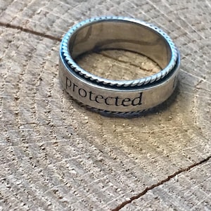 Op de afbeelding: Een zilveren ring met het woord "protected" gegraveerd op de band. De ring heeft een gevlochten touwontwerp rond de randen en ligt op een houten oppervlak.