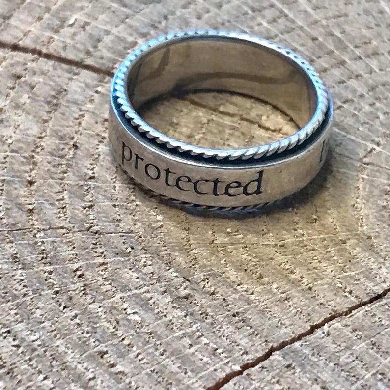 Op de afbeelding: Een zilveren ring met het woord "protected" gegraveerd op de band. De ring heeft een gevlochten touwontwerp rond de randen en ligt op een houten oppervlak.