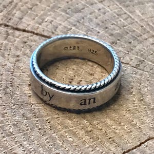 Op de afbeelding: Een zilveren ring met een gevlochten rand en de woorden "by an" gegraveerd op de band. De ring heeft de markeringen "&copy;t&t. 925" aan de binnenkant van de band. De ring ligt op een rustiek houten oppervlak.