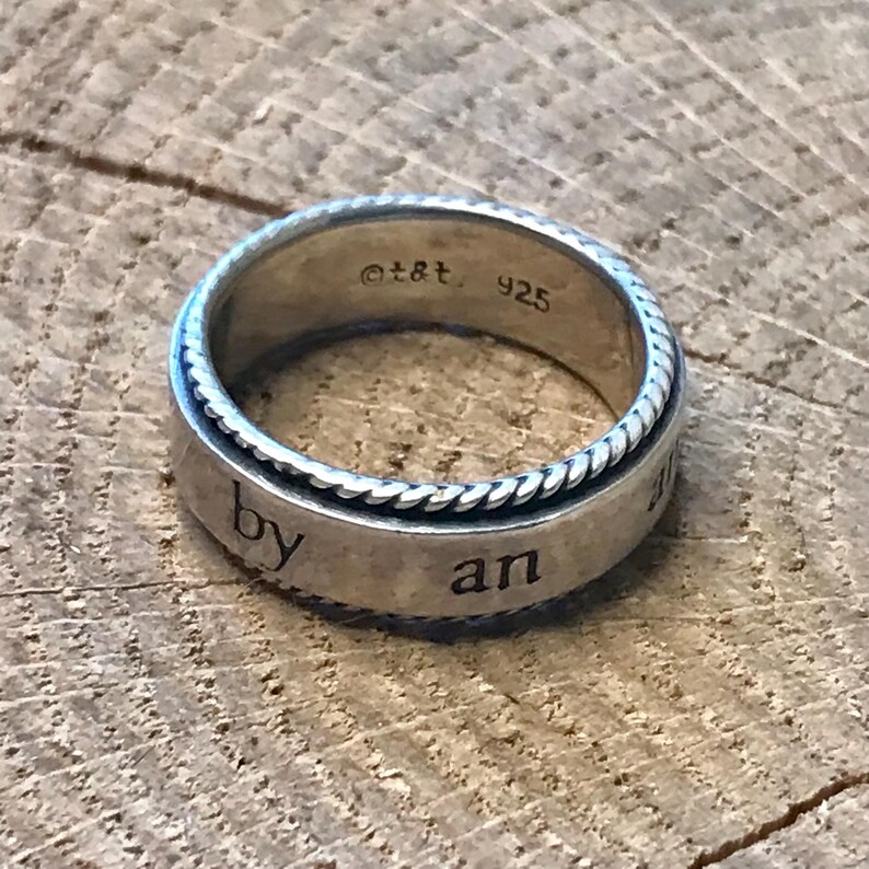 Op de afbeelding: Een zilveren ring met een gevlochten rand en de woorden "by an" gegraveerd op de band. De ring heeft de markeringen "&copy;t&t. 925" aan de binnenkant van de band. De ring ligt op een rustiek houten oppervlak.