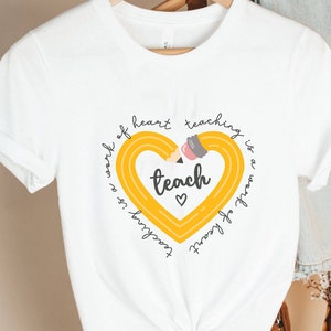 May include: White t-shirt with a yellow pencil heart graphic and the text "Teaching is a work of heart. Teaching is a work of heart. Teach."