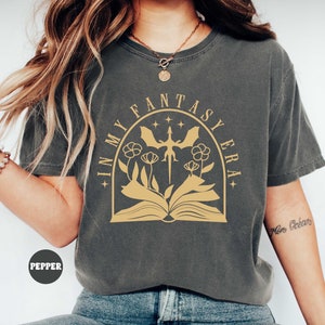 May include: A gray t-shirt with the text "In My Fantasy Era" in a gold arch with a dragon and flowers. The text is in a vintage font.
