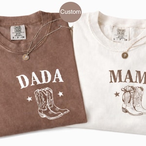 Könnte beinhalten: Zwei T-Shirts, eines in Espresso und eines in Elfenbein, mit den Aufdrucken "DADA" und "MAMA". Jedes Shirt hat eine Grafik von Cowboy-Stiefeln und Sternen. Beide Shirts haben eine Halskette.