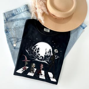 May include: Black t-shirt featuring a graphic of characters crossing a street with a full moon and spooky buildings in the background. The shirt is paired with light blue jeans and a tan hat.