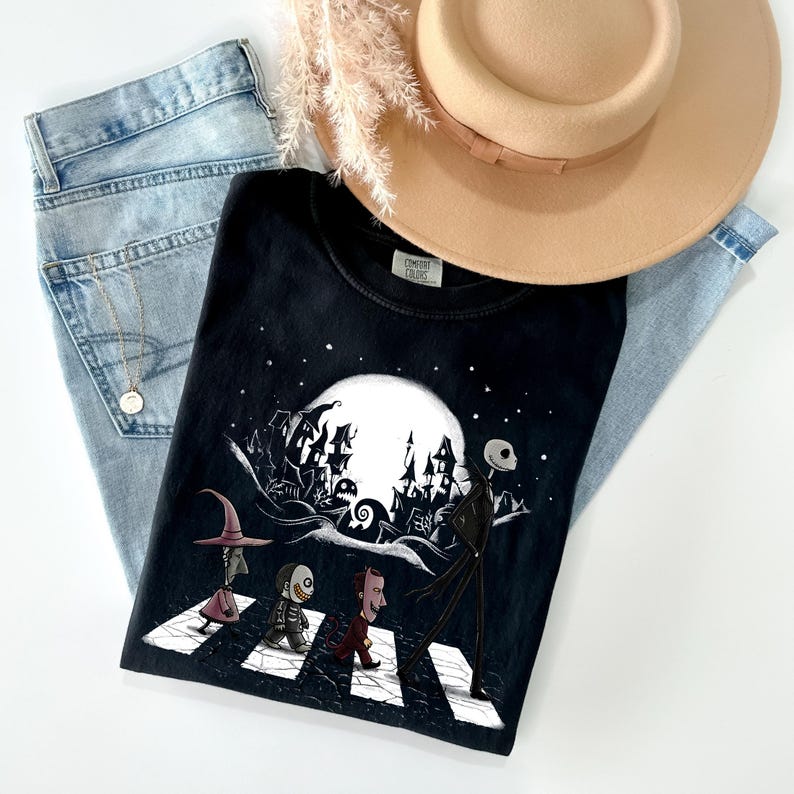 May include: Black t-shirt featuring a graphic of characters crossing a street with a full moon and spooky buildings in the background. The shirt is paired with light blue jeans and a tan hat.