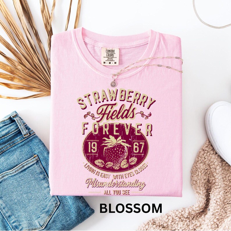 a pink t-shirt with a design that says "STRAWBERRY FIELDS FOREVER" and "LIVING IS EASY WITH EYES CLOSED" along with a strawberry graphic. The t-shirt is displayed on a white background, accompanied by a pair of blue jeans, a white scarf, and a white mug.