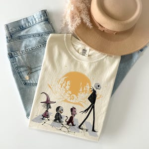 May include: Cream-colored t-shirt featuring a cartoon illustration of characters walking in front of a yellow sun and a castle silhouette. The shirt is paired with light blue jeans, a gold necklace, and a tan hat.