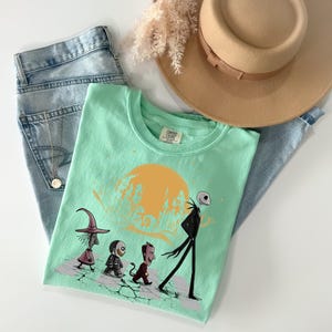 May include: A light green t-shirt featuring a graphic of characters walking in front of a yellow moon. The shirt is paired with light blue jeans and a tan hat. A gold necklace is visible on the jeans.
