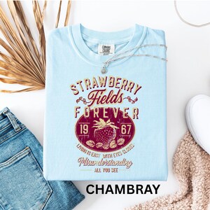 a light blue t-shirt with a design that says "STRAWBERRY FIELDS FOREVER" and "LOVING IS EASY WITH EYES CLOSED" printed on it. The t-shirt is laid out on a white surface, accompanied by a pair of jeans, a necklace, and a beige sweater.
