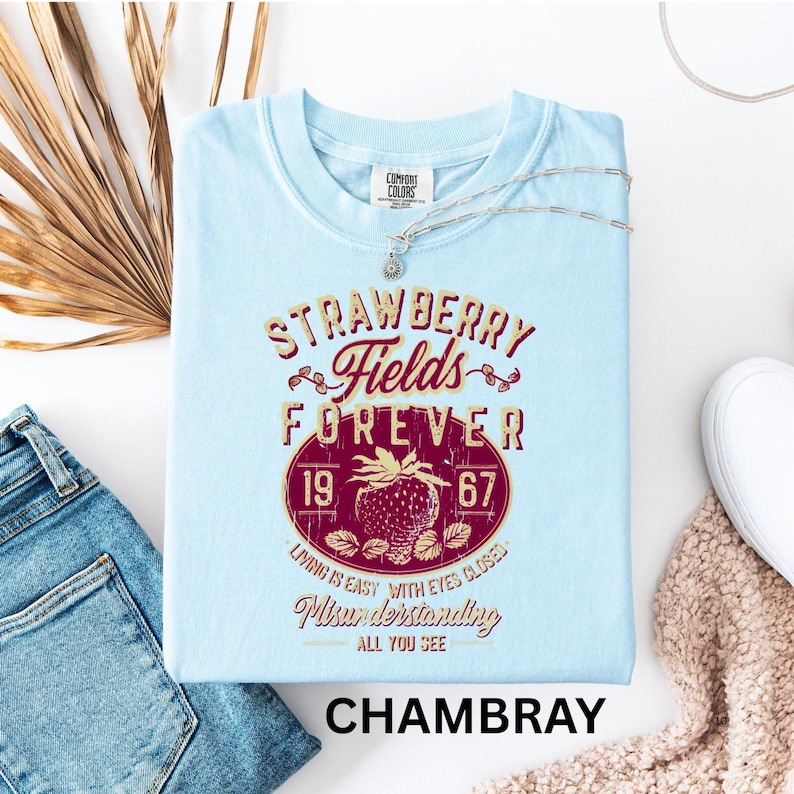 a light blue t-shirt with a design that says "STRAWBERRY FIELDS FOREVER" and "LOVING IS EASY WITH EYES CLOSED" printed on it. The t-shirt is laid out on a white surface, accompanied by a pair of jeans, a necklace, and a beige sweater.