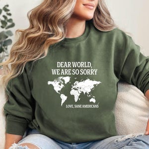May include: Olive green sweatshirt with the text "DEAR WORLD, WE ARE SO SORRY" above a white world map graphic. Below the map, the text reads "LOVE, SANE AMERICANS". The sweatshirt is a casual style.