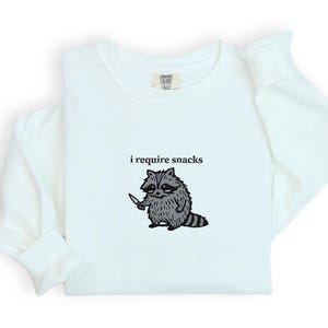 May include: White long-sleeve shirt with the text "i require snacks" above a cartoon raccoon holding a knife. The raccoon is gray and black with a striped tail. The shirt is a light color.