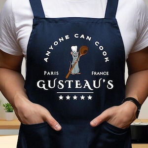 Könnte beinhalten: Marineblaue Schürze mit dem weißen Text "ANYONE CAN COOK" und "GUSTEAU'S PARIS FRANCE". In der Mitte ist eine Cartoon-Ratte mit einem Holzlöffel abgebildet. Die Schürze hat zwei Taschen und schwarze Träger.