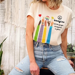 A woman wearing a white t-shirt with a peace sign and colorful hand-drawn figures, sitting in front of a white wooden wall with plants.