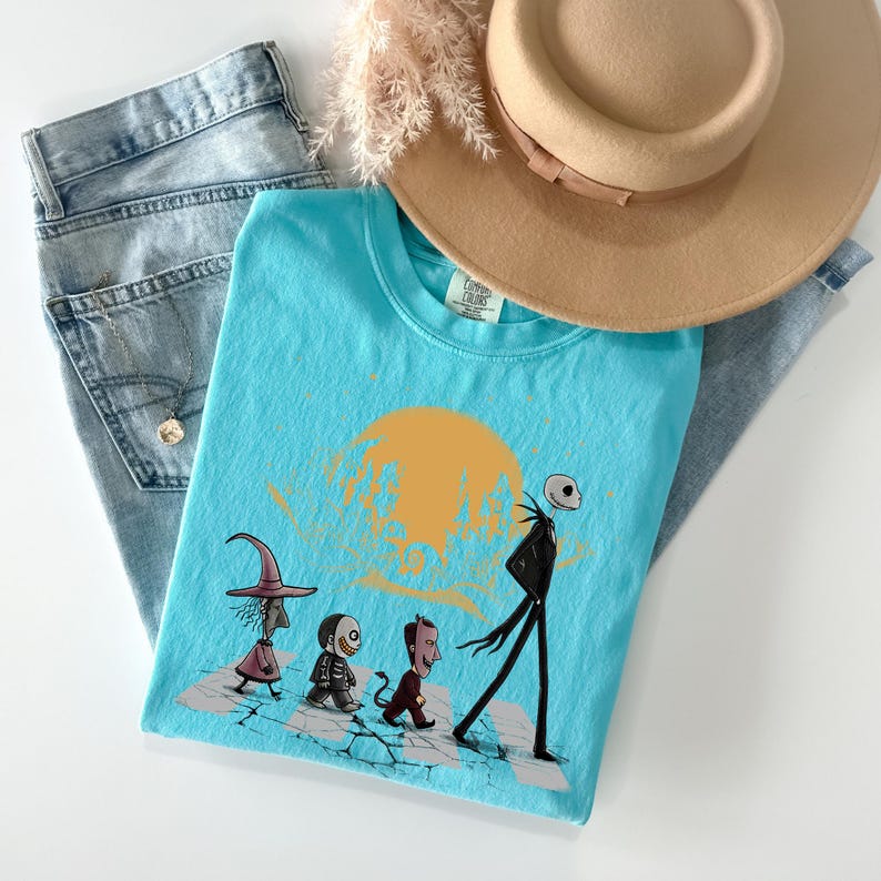 May include: A light blue t-shirt featuring a cartoon illustration of characters walking across a crosswalk, with a yellow sun in the background. The shirt is paired with blue jeans and a tan hat.