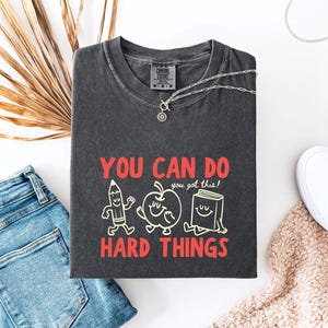 May include: A charcoal gray t-shirt featuring a graphic with a smiling pencil, apple, and book. The text reads "YOU CAN DO HARD THINGS" in red letters, with "you got this!" in smaller white script. The shirt has a crew neck and a Comfort Colors label.
