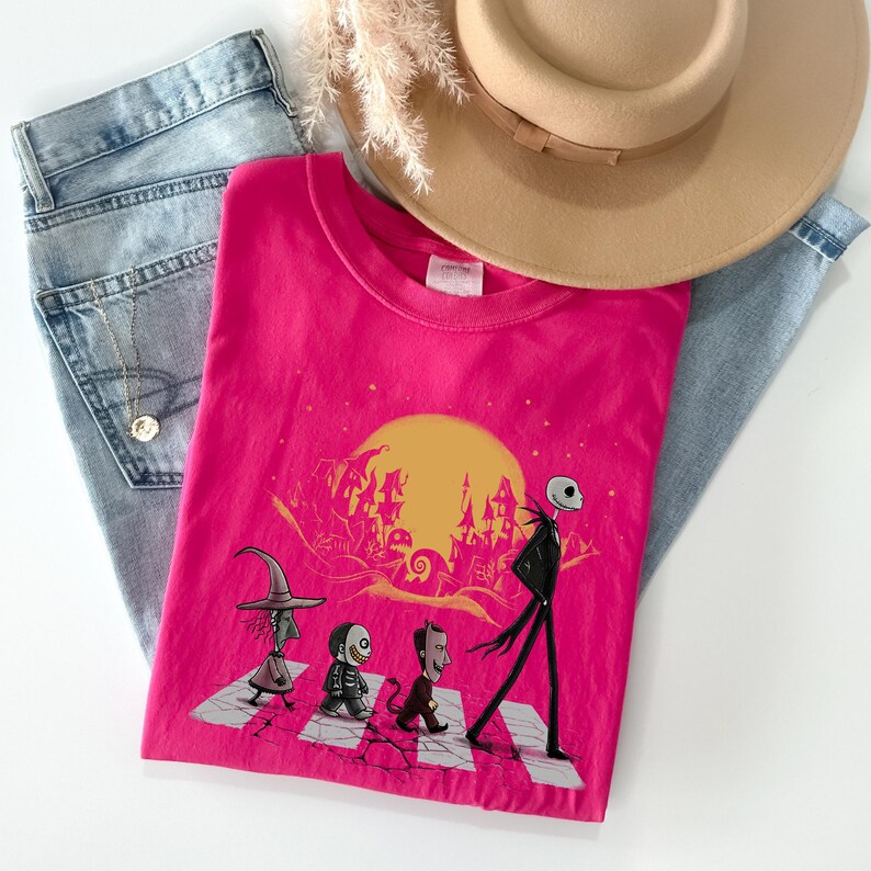 May include: A hot pink t-shirt featuring a graphic of characters crossing a street with a yellow moon and a spooky town in the background. The shirt is laid on top of blue jeans and a tan hat.