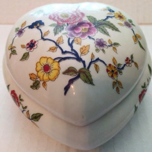 May include: A white porcelain heart-shaped trinket box with a floral design. The flowers are yellow, pink, red, and purple with green leaves and blue stems.