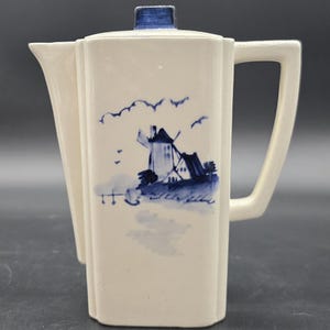 May include: Cream-colored ceramic pitcher with a blue windmill and landscape design. The pitcher has a rectangular shape with a handle and a spout. The lid is blue. The design includes a windmill, birds, and a field.