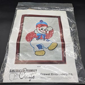 May include: A vintage "American Family Crafts" crewel embroidery kit. The kit features a framed image of a smiling cartoon character in a red and white checkered shirt, blue hat, and yellow shoes. The image is in a plastic bag.