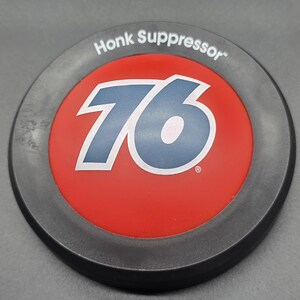 May include: A round, gray and black "Honk Suppressor" emblem. The center features a red circle with the number "76" in blue and white. The emblem has a distressed, weathered appearance.