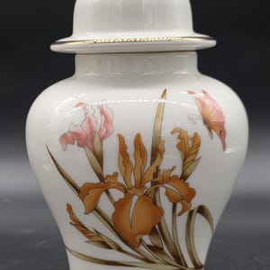 May include: White ceramic jar with a lid, featuring a floral design of orange and pink irises and a butterfly. The jar has gold trim and a gold finial on the lid. A decorative piece for the home.