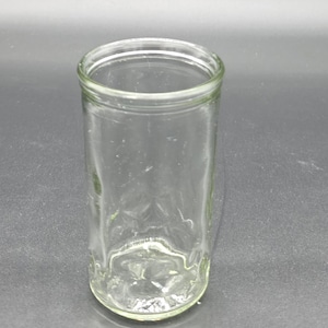 May include: A clear, cylindrical glass jar with a wide opening. The jar is empty and sits on a dark surface. The glass has a slight green tint.
