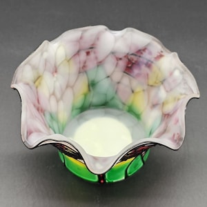 May include: A decorative glass bowl with a ruffled rim. The bowl has a white, pink, yellow, and green pattern inside. The base is green with black and red accents. The bowl has a smooth, glossy finish.
