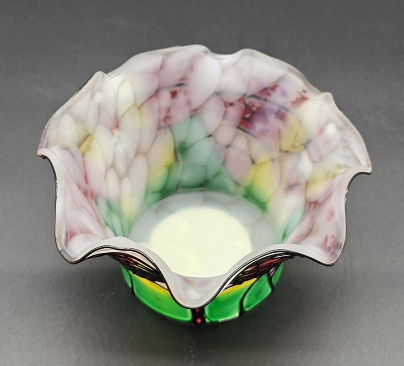 May include: A decorative glass bowl with a ruffled rim. The bowl has a white, pink, yellow, and green pattern inside. The base is green with black and red accents. The bowl has a smooth, glossy finish.