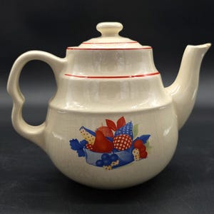 May include: Cream ceramic teapot with a fruit basket design. The teapot has a curved handle, a spout, and a lid with a small knob. Red trim accents the lid and body. The fruit basket features red, blue, and yellow fruits.