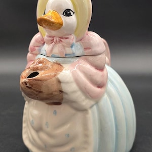 Puede incluir: Una azucarera o jarra de crema con forma de pato de cerámica. El pato lleva un gorro amarillo, un lazo rosa y un vestido azul claro. La jarra de crema tiene un pico en forma de cesta marrón.