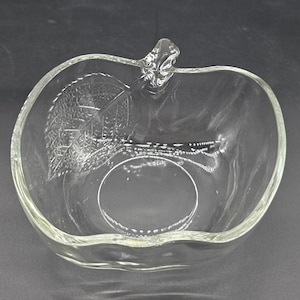 May include: Clear glass apple-shaped bowl with a stem and leaf design. The bowl has a textured leaf pattern on one side and a smooth, oval base. The bowl is transparent, allowing light to pass through.