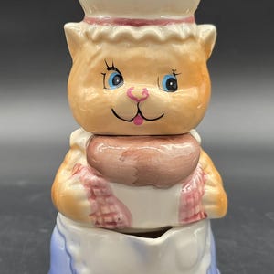 Puede incluir: Un recipiente de cerámica con forma de gato con gorro y delantal de chef. El gato tiene pelaje naranja, ojos azules y nariz rosa. El delantal es blanco con ribete azul, y el recipiente está en tres secciones apiladas.