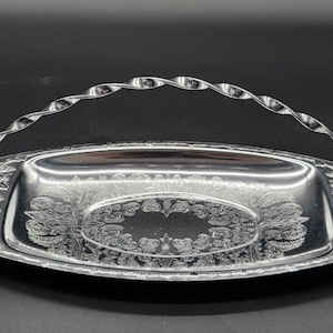 May include: A silver-colored, rectangular serving tray with a decorative floral pattern and a twisted handle. The tray is made of metal and has an oval design in the center. The handle is attached to the sides of the tray.