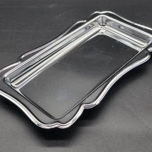 Puede incluir: Una bandeja rectangular plateada brillante con un borde festoneado. La bandeja tiene un borde elevado y una superficie lisa y reflectante. El diseño es simple y elegante, adecuado para servir o exhibir.
