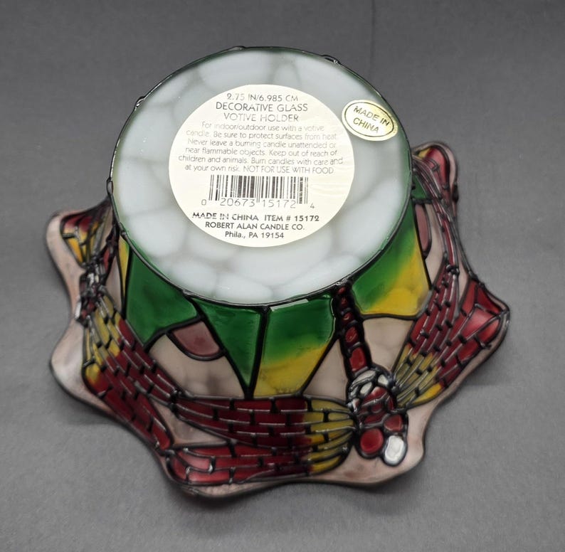 May include: Decorative glass votive holder with a dragonfly design. The holder is 6.985 cm in diameter and features red, yellow, and green stained glass. The base is shaped like a flower.