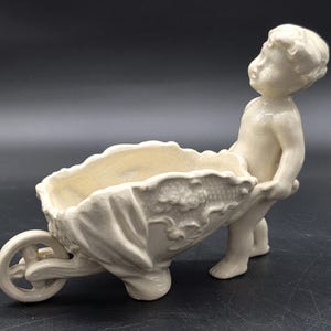 May include: A cream-colored ceramic planter shaped like a wheelbarrow being pushed by a cherubic figure. The wheelbarrow has a textured floral design and a shallow basin for plants. The figure is in a standing position, with arms outstretched.