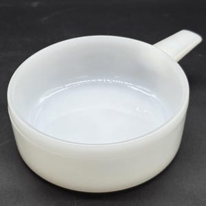 May include: A white ceramic serving bowl with a handle. The bowl is round and has a smooth, glossy finish. The handle is rectangular and extends from the side. The bowl is empty and likely for serving soups or side dishes.