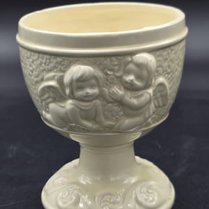 May include: Cream-colored ceramic goblet with a pedestal base. The goblet features embossed cherub figures and floral designs. The cup is approximately 5 inches tall and 4 inches wide.