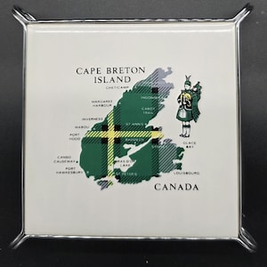 Könnte beinhalten: Quadratischer Untersetzer mit einer Karte von Cape Breton Island, Kanada, in grün-gelbem Karomuster. Der Untersetzer hat einen weißen Hintergrund mit Ortsnamen und einer Zeichnung eines Dudelsackspielers. Er ist in silbernem Metall gerahmt.