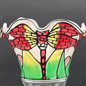 May include: A decorative glass bowl with a dragonfly design. The bowl has a white, scalloped rim and a colourful dragonfly motif in red, yellow, and green. The dragonfly's wings have a brick-like appearance.