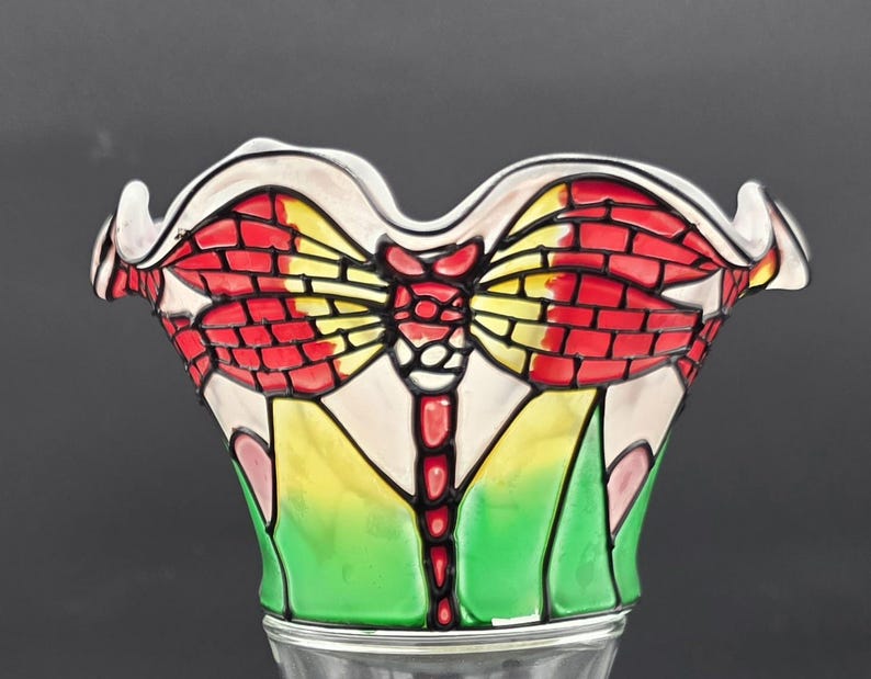 May include: A decorative glass bowl with a dragonfly design. The bowl has a white, scalloped rim and a colourful dragonfly motif in red, yellow, and green. The dragonfly's wings have a brick-like appearance.